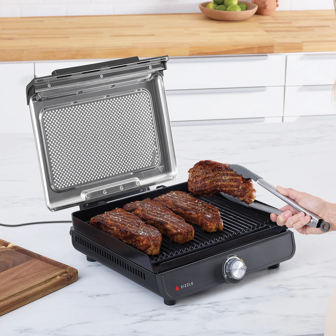 Ninja-GR101UK-InUse-Kitchen-OnIsland-HighHeatGrilling-4Steaks-Kitchen-Woman-RGB